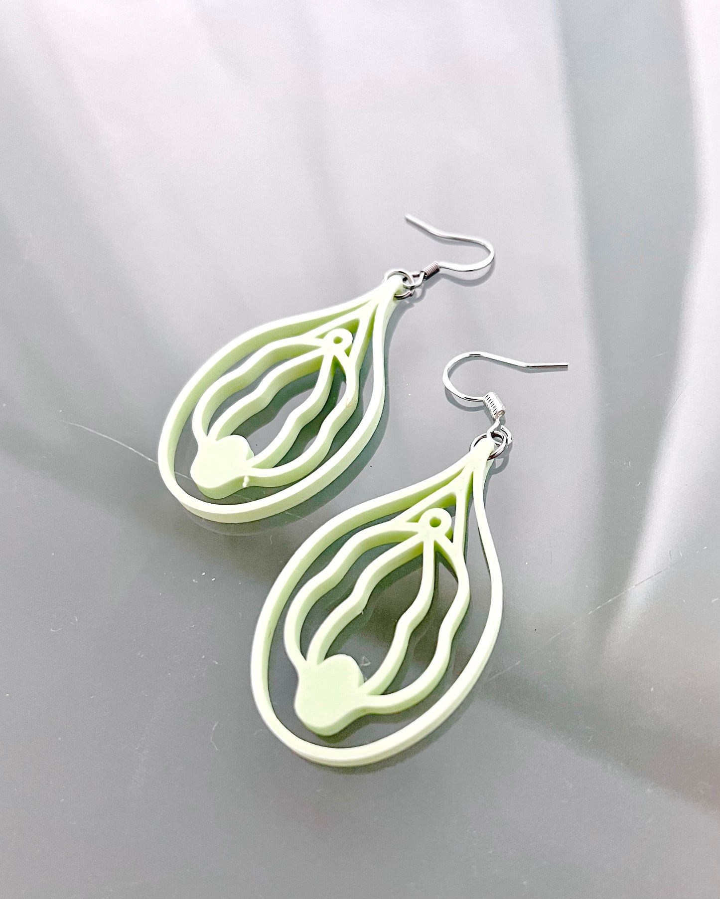 Light green vulva shaped earrings on a reflective gray surface