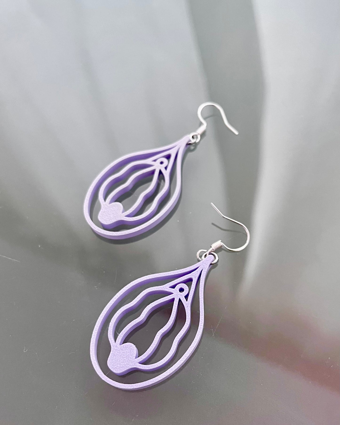 Pair of purple or lilac vulva shaped earrings on a reflective gray surface