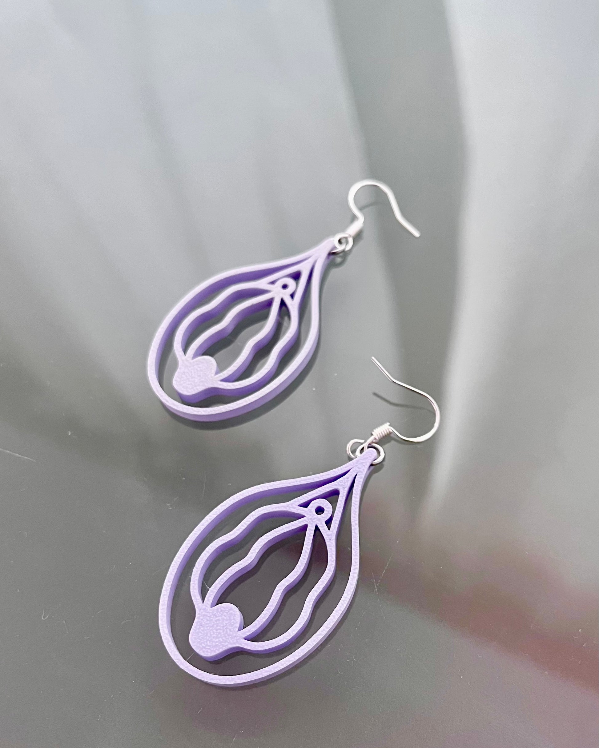 Pair of purple or lilac vulva shaped earrings on a reflective gray surface