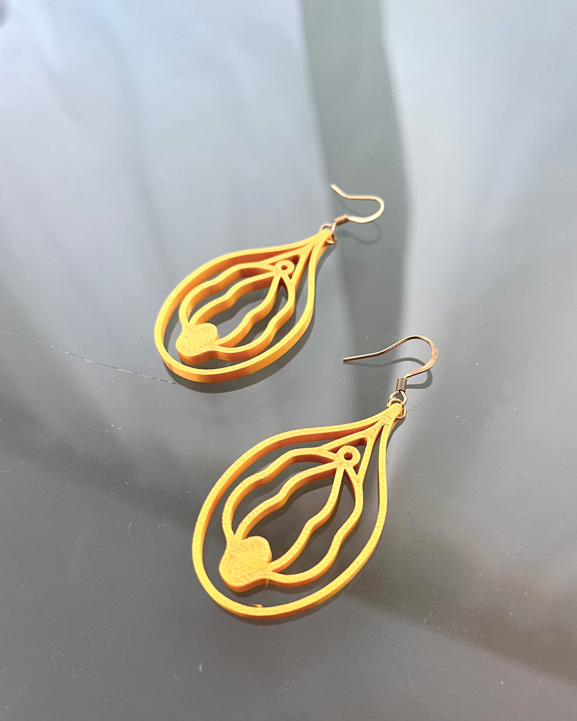 Gold vulva shaped earrings on a reflective gray surface